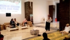 Woman in a hijab speaking into a microphone at a conference panel; three people seated on beige couches with drinks, a projection screen behind them.