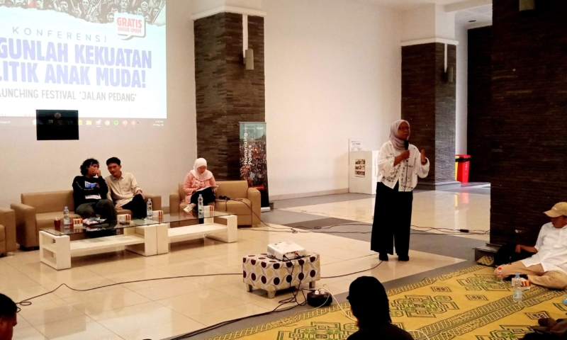 Woman in a hijab speaking into a microphone at a conference panel; three people seated on beige couches with drinks, a projection screen behind them.
