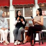 Three women sit on stools on a red carpet, each holding a microphone during a panel discussion outside a glass-walled venue with red awnings.