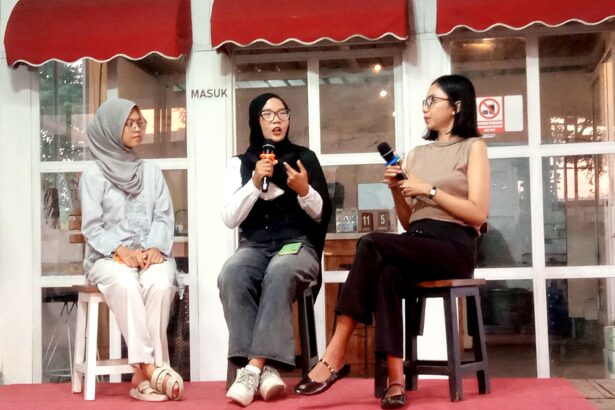 Three women sit on stools on a red carpet, each holding a microphone during a panel discussion outside a glass-walled venue with red awnings.
