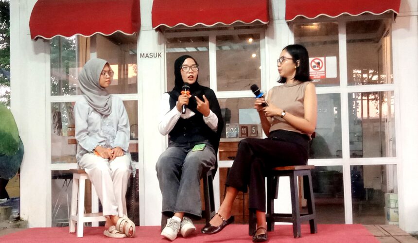 Three women sit on stools on a red carpet, each holding a microphone during a panel discussion outside a glass-walled venue with red awnings.