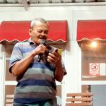 Man in a striped polo speaks into a handheld microphone outside a building with red awnings behind him.