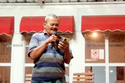 Man in a striped polo speaks into a handheld microphone outside a building with red awnings behind him.