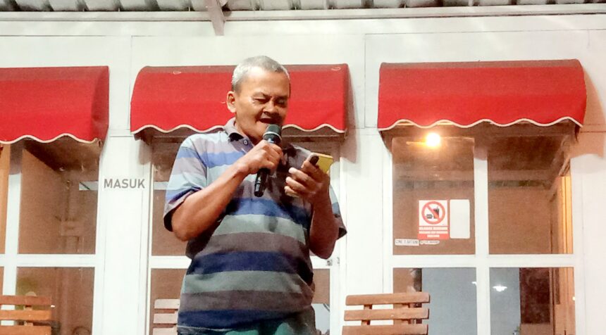Man in a striped polo speaks into a handheld microphone outside a building with red awnings behind him.