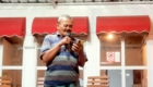 Man in a striped polo speaks into a handheld microphone outside a building with red awnings behind him.
