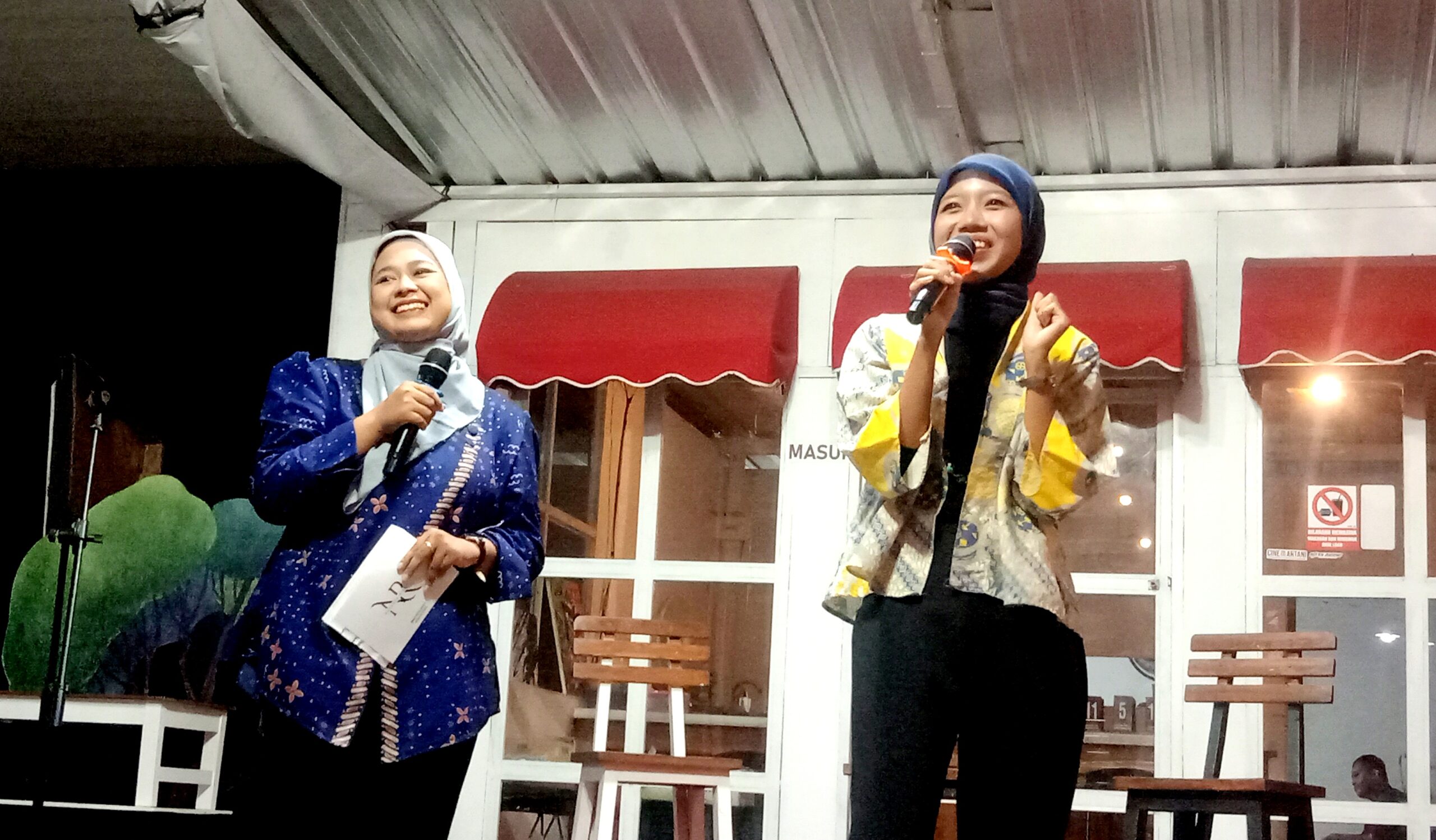 Two women wearing hijabs on a stage, each holding a microphone and smiling during a performance or presentation.
