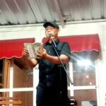 Man in a black shirt and cap reading from a booklet into a microphone on a small stage indoors.