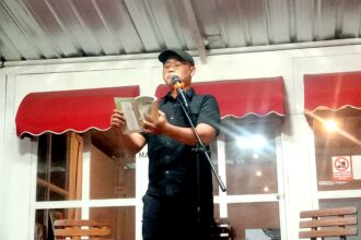 Man in a black shirt and cap reading from a booklet into a microphone on a small stage indoors.