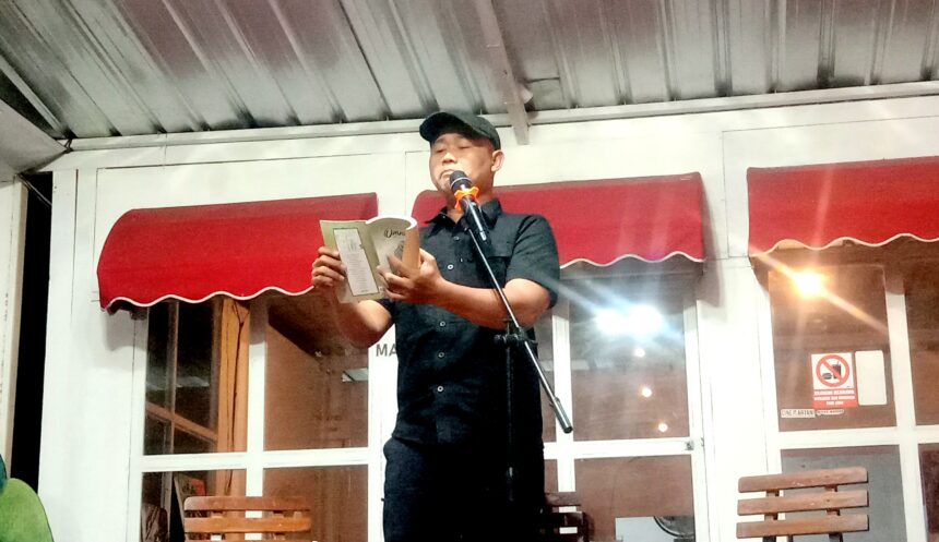 Man in a black shirt and cap reading from a booklet into a microphone on a small stage indoors.