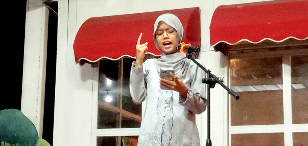 Woman wearing a light gray hijab and dress speaks into a microphone on stage, gesturing with one finger while holding a phone.