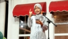 Woman wearing a light gray hijab and dress speaks into a microphone on stage, gesturing with one finger while holding a phone.