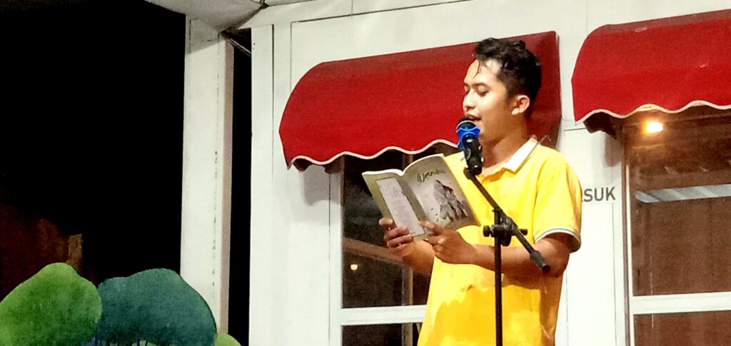 Young man in a yellow shirt reads from a magazine into a microphone on a small stage with a red awning backdrop.