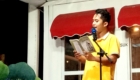Young man in a yellow shirt reads from a magazine into a microphone on a small stage with a red awning backdrop.