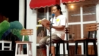 Man seated on a stool reads aloud from a book into a microphone on a small stage, red awning and white-framed windows behind him.