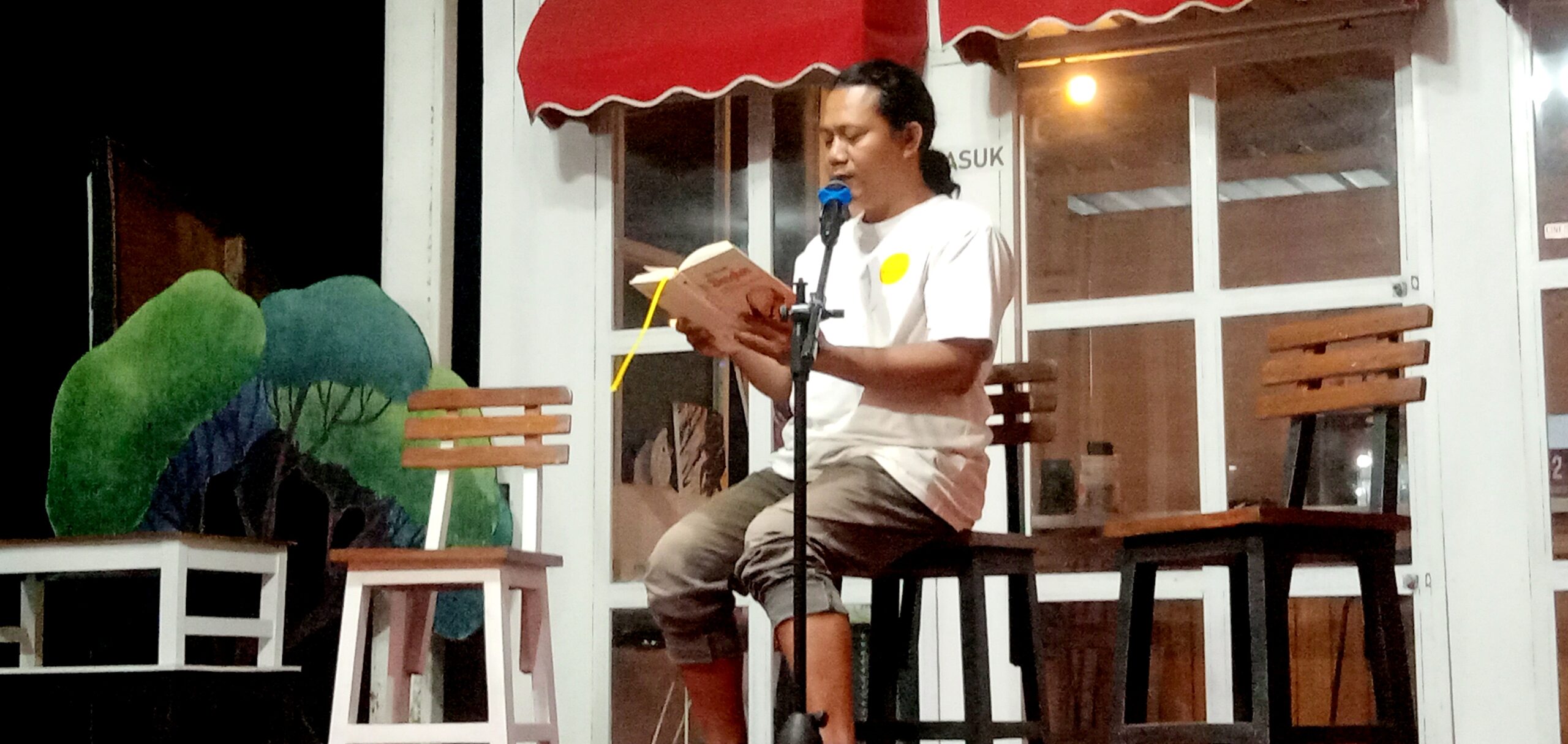 Man seated on a stool reads aloud from a book into a microphone on a small stage, red awning and white-framed windows behind him.