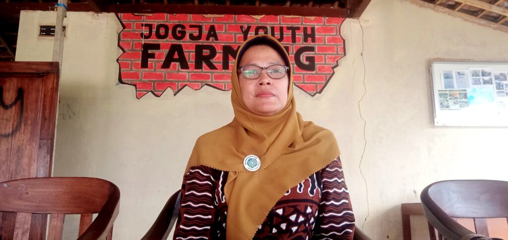 IMG_20260612_155611 - Mabur.co Portrait of a woman in a mustard hijab and glasses, seated indoors with a red brick mural that reads'JOGJA YOUTH FARMING' behind her.