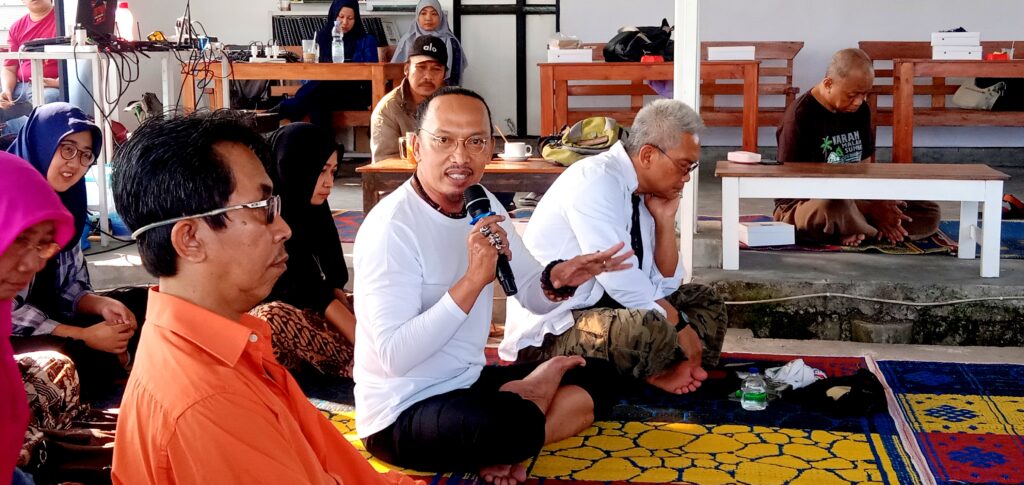 Man in a white shirt speaks into a handheld microphone while seated on a colorful rug, addressing a group indoors.