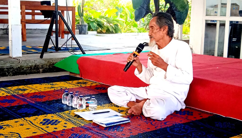 Elderly man in white robes sitting cross‑legged on a colorful rug, speaking into a microphone in an outdoor space with plants nearby and water bottles on the ground.
