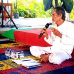 Elderly man in white robes sitting cross‑legged on a colorful rug, speaking into a microphone in an outdoor space with plants nearby and water bottles on the ground.