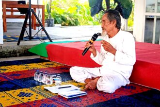 Elderly man in white robes sitting cross‑legged on a colorful rug, speaking into a microphone in an outdoor space with plants nearby and water bottles on the ground.