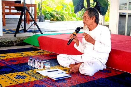 Elderly man in white robes sitting cross‑legged on a colorful rug, speaking into a microphone in an outdoor space with plants nearby and water bottles on the ground.
