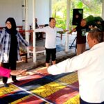 People of diverse ages form a circle with arms extended, participating in a group stretch on a colorful mat.