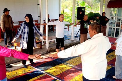 People of diverse ages form a circle with arms extended, participating in a group stretch on a colorful mat.