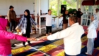 People of diverse ages form a circle with arms extended, participating in a group stretch on a colorful mat.