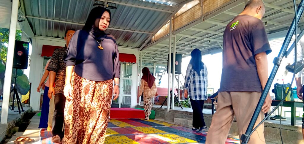 Woman in a dark blouse and batik skirt stands on a colorful patterned stage rug at an outdoor event under a metal pavilion door frame.