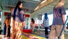 Woman in a dark blouse and batik skirt stands on a colorful patterned stage rug at an outdoor event under a metal pavilion door frame.