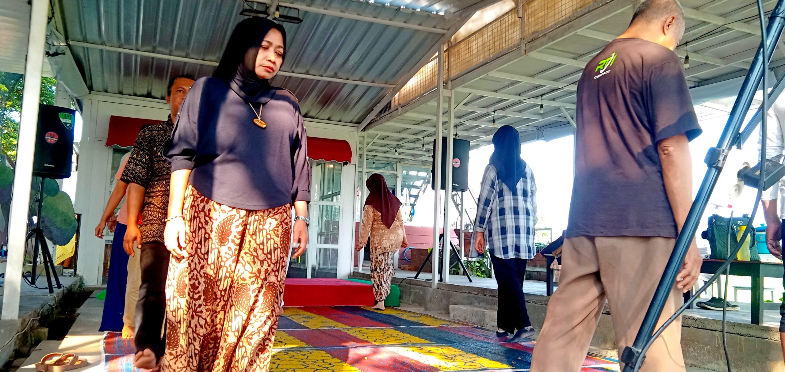 Woman in a dark blouse and batik skirt stands on a colorful patterned stage rug at an outdoor event under a metal pavilion door frame.