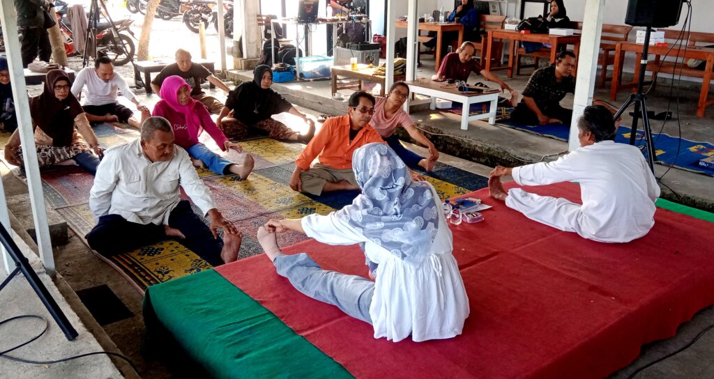 Group of people seated on colorful mats in a semicircle, performing leg stretches as an instructor in white robes leads from a red platform.
