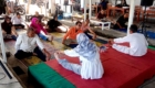 Group of people seated on colorful mats in a semicircle, performing leg stretches as an instructor in white robes leads from a red platform.