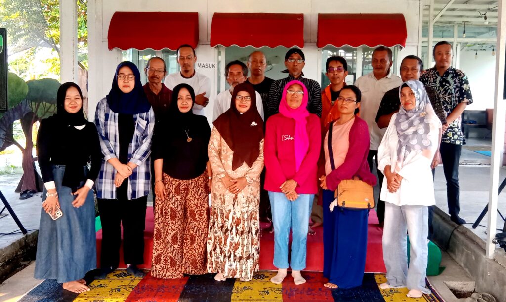Group portrait of about 14 adults standing shoulder-to-shoulder on a colorful carpet under red canopy awnings, smiling at the camera.