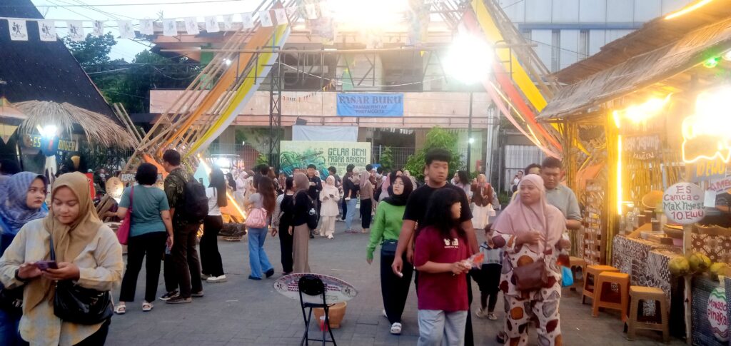 Crowded outdoor market street with stalls, colorful banners, and people browsing at dusk/evening.
