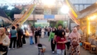 Crowded outdoor market street with stalls, colorful banners, and people browsing at dusk/evening.
