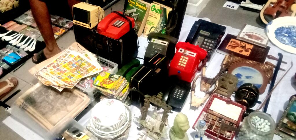 Table full of vintage electronics and knickknacks at a flea market stall, including red payphone, old phones, magazines, and dishes.