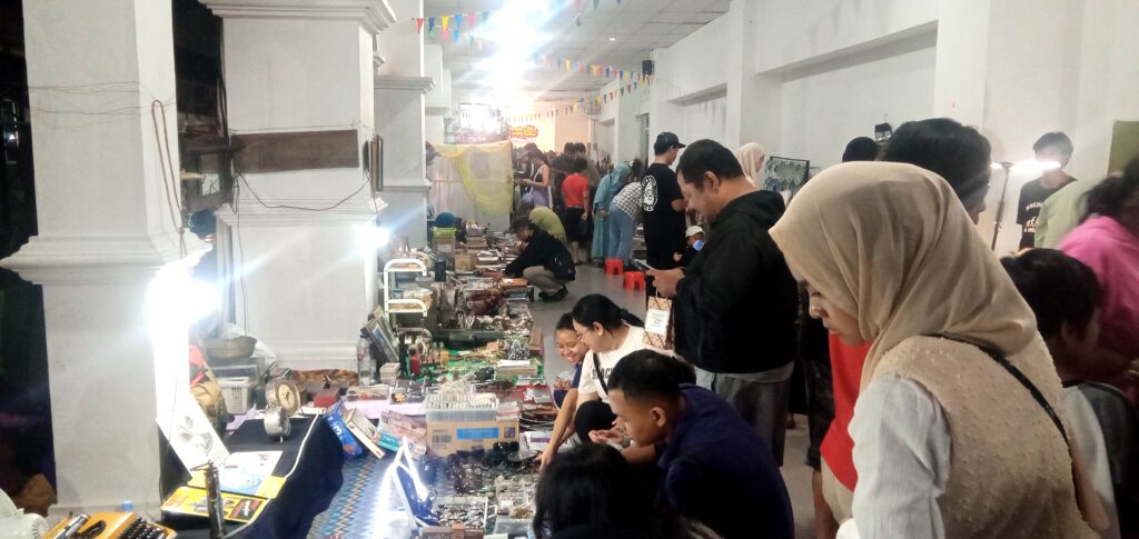 Crowded indoor market with vendor stalls lining a long hallway, shoppers browsing rows of small goods beneath colorful bunting.