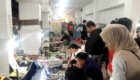 Crowded indoor market with vendor stalls lining a long hallway, shoppers browsing rows of small goods beneath colorful bunting.