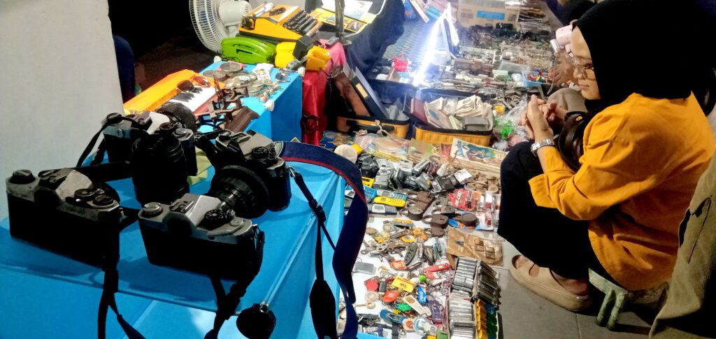 Two people browse a busy flea-market stall with cameras and vintage gadgets on blue tables.