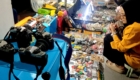 Two people browse a busy flea-market stall with cameras and vintage gadgets on blue tables.