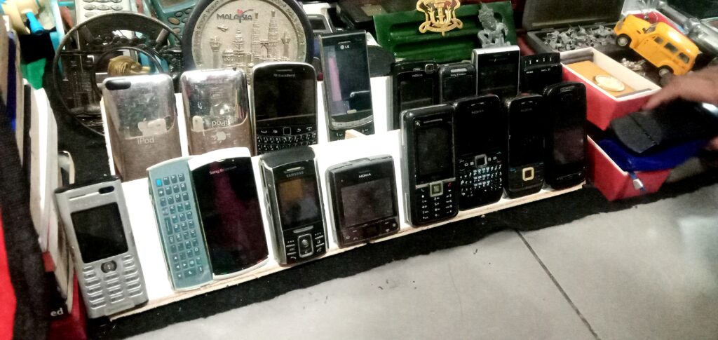 Shelf displaying vintage mobile phones (Nokia, Sony Ericsson) and iPods lined up for sale.