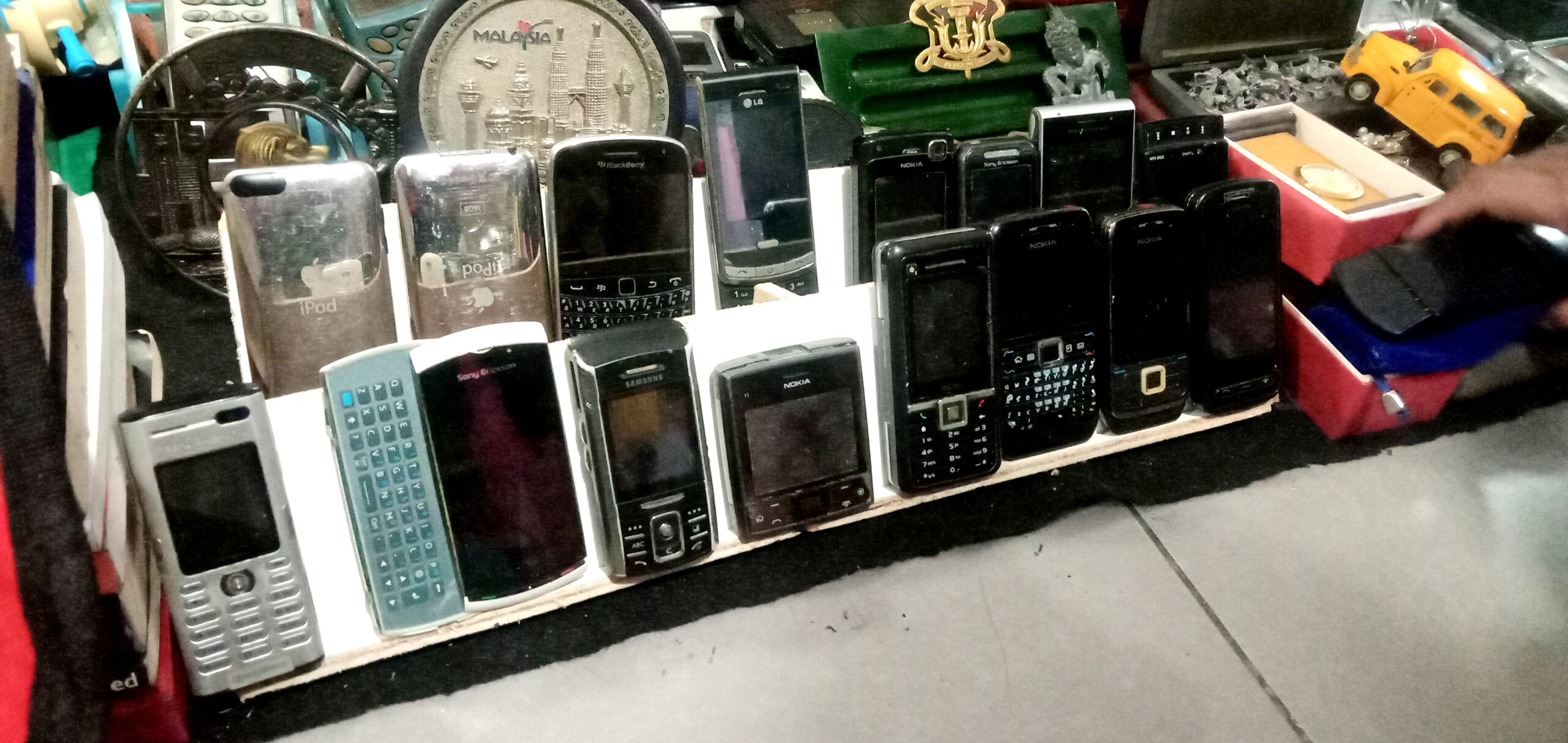 Shelf displaying vintage mobile phones (Nokia, Sony Ericsson) and iPods lined up for sale.