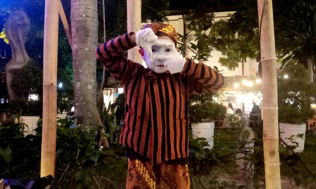 Mime performer in orange-and-black striped shirt, white face makeup, and patterned headscarf posing with hands framing eyes outdoors at night.