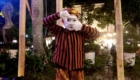 Mime performer in orange-and-black striped shirt, white face makeup, and patterned headscarf posing with hands framing eyes outdoors at night.