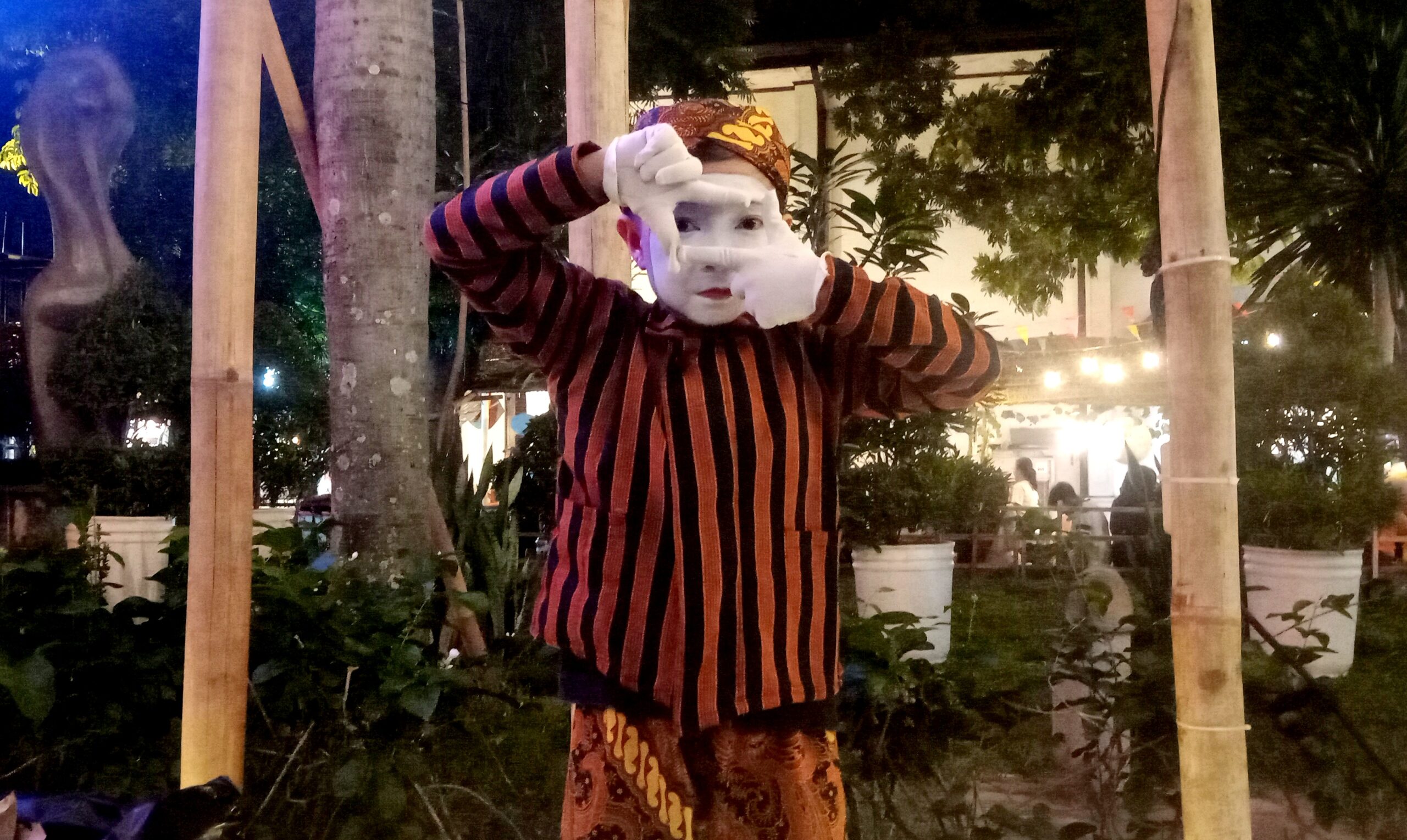Mime performer in orange-and-black striped shirt, white face makeup, and patterned headscarf posing with hands framing eyes outdoors at night.