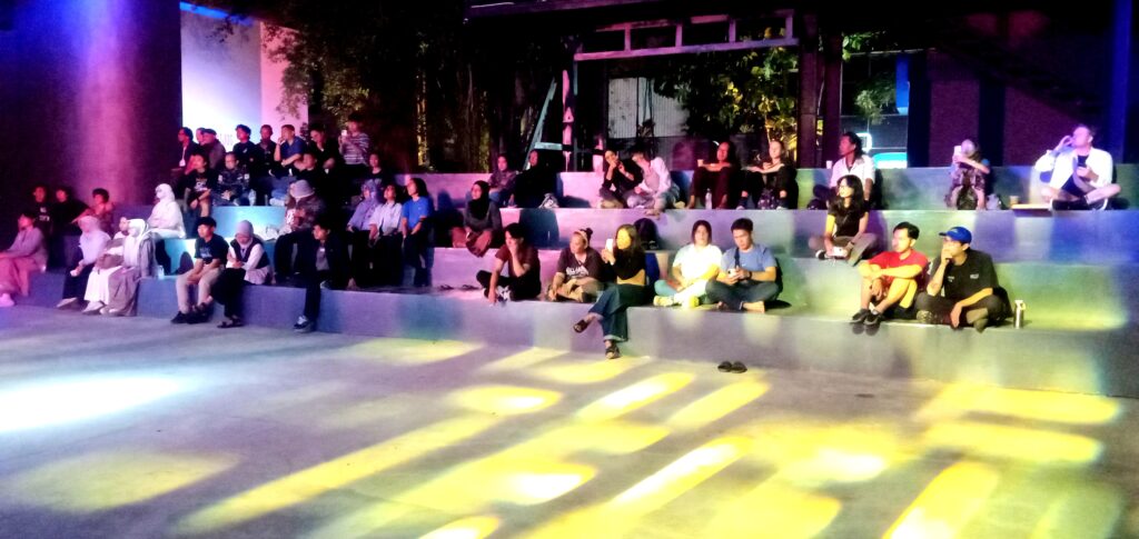 Audience seated on colorful, tiered bleachers watching a nighttime outdoor performance under stage lighting, with people chatting and observing the show.