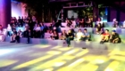 Audience seated on colorful, tiered bleachers watching a nighttime outdoor performance under stage lighting, with people chatting and observing the show.