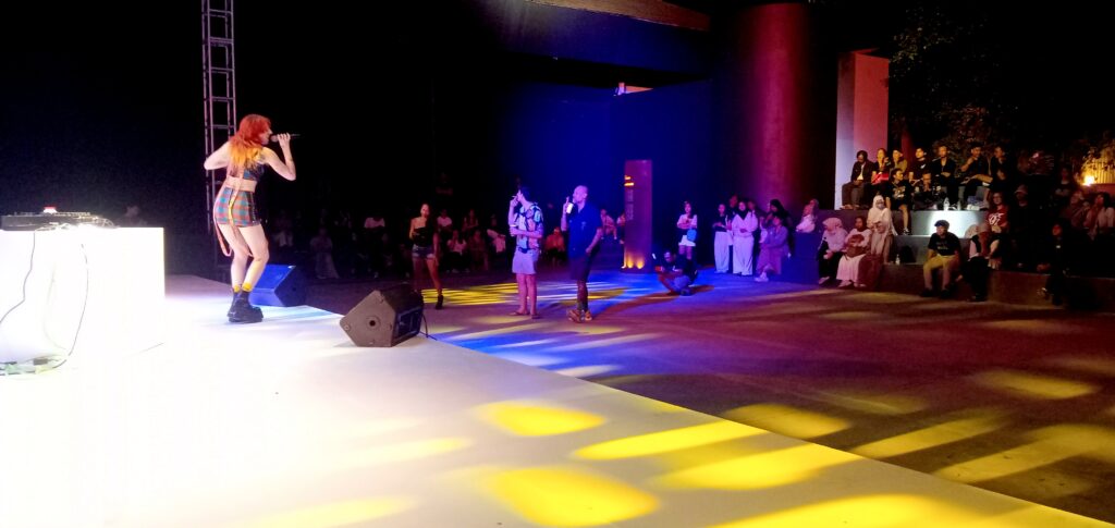 Female singer on stage using a microphone, wearing a colorful plaid outfit, performing to a night-time audience.