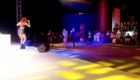 Female singer on stage using a microphone, wearing a colorful plaid outfit, performing to a night-time audience.
